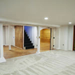 Bright basement space with white support columns, a carpeted main area, and a wooden arched doorway beside a short black staircase.
