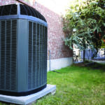 Outdoor air conditioner condenser unit on a concrete pad beside a brick wall and green lawn.