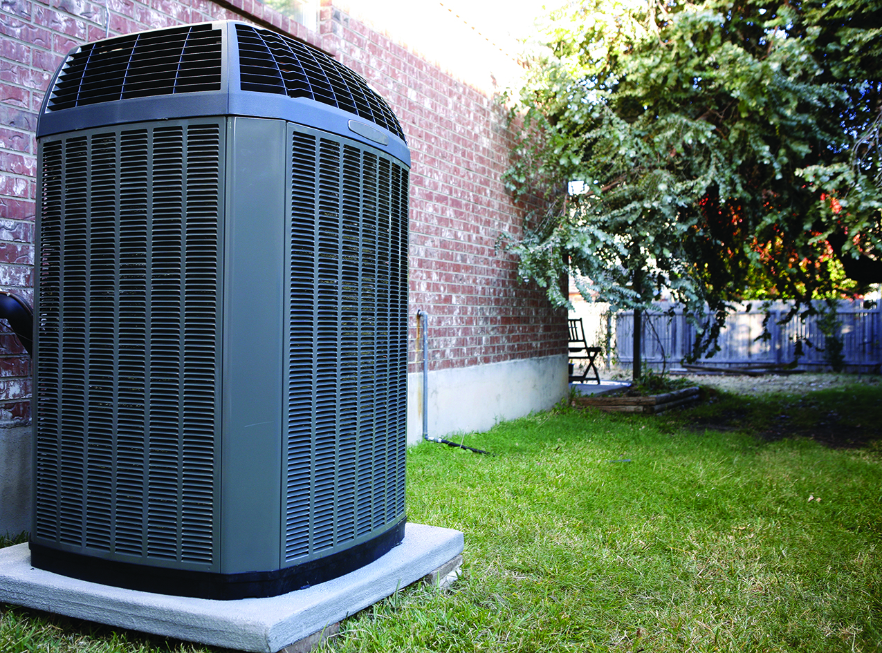 Outdoor air conditioner condenser unit on a concrete pad beside a brick wall and green lawn.