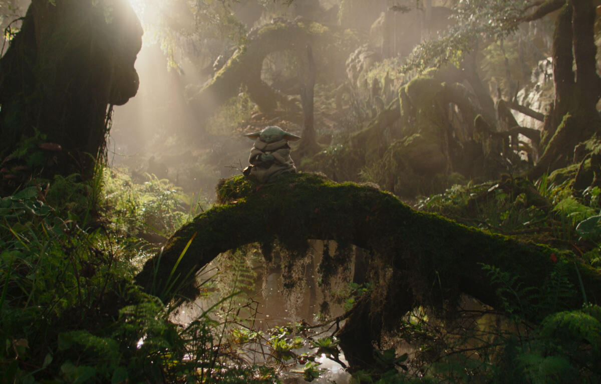 Grogu (Baby Yoda) statue sits on a mossy log in a sunlit, misty forest.