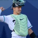 Baseball catcher in light mint-green chest protector and mask, mid-throw against a blue geometric backdrop.