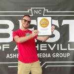 Smiling man in a red shirt holds a 2025 winner plaque in front of a 'Best of Asheville' banner backdrop.