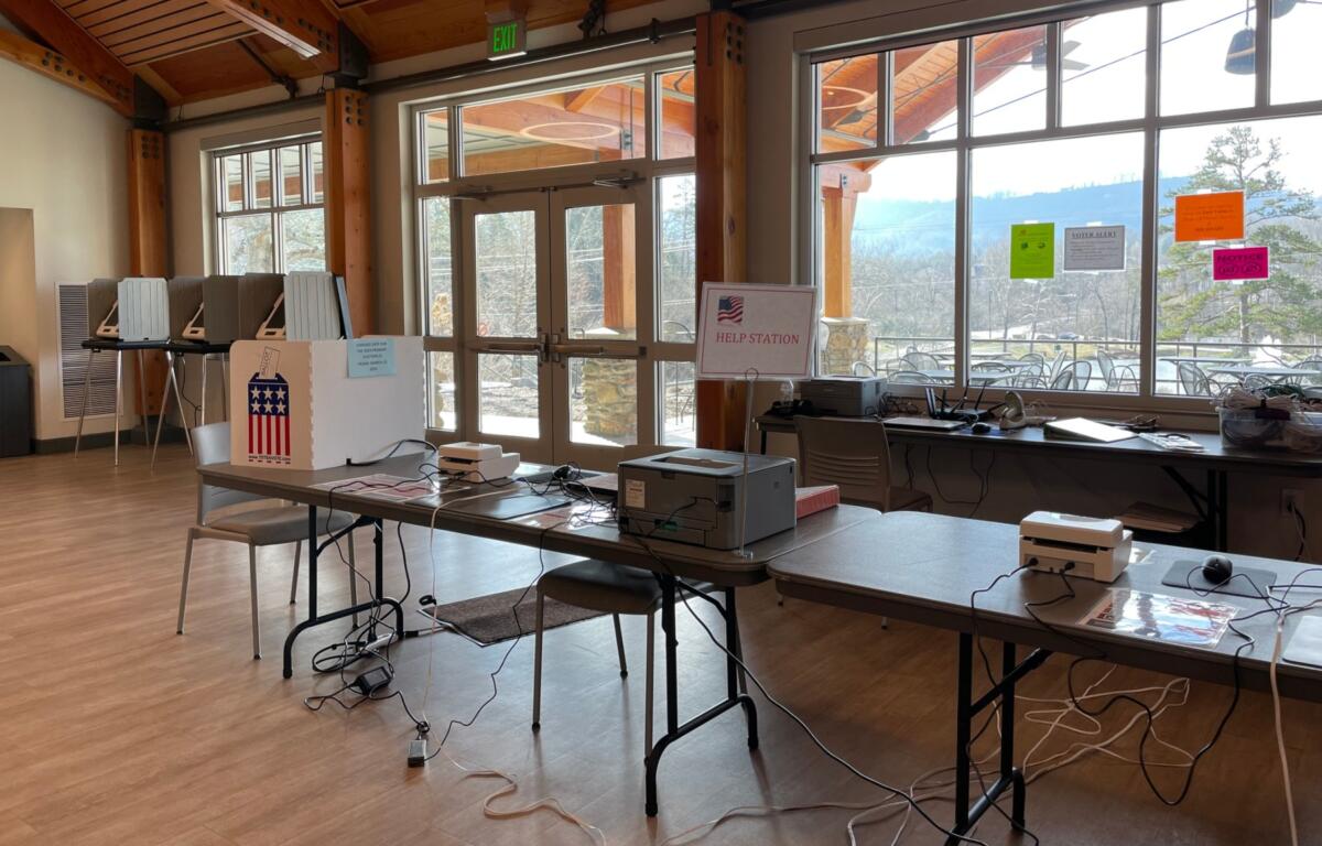 Election help station set up in a bright community room with voting equipment, a Help Station sign, and tangled cables on tables.