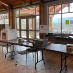 Election help station set up in a bright community room with voting equipment, a Help Station sign, and tangled cables on tables.