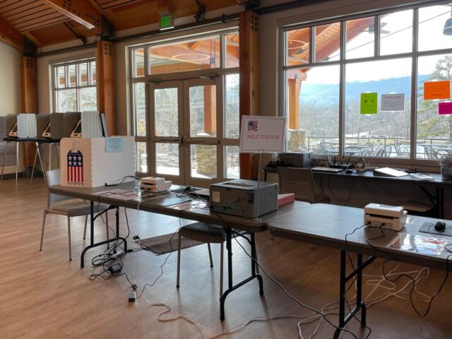 Election help station set up in a bright community room with voting equipment, a Help Station sign, and tangled cables on tables.