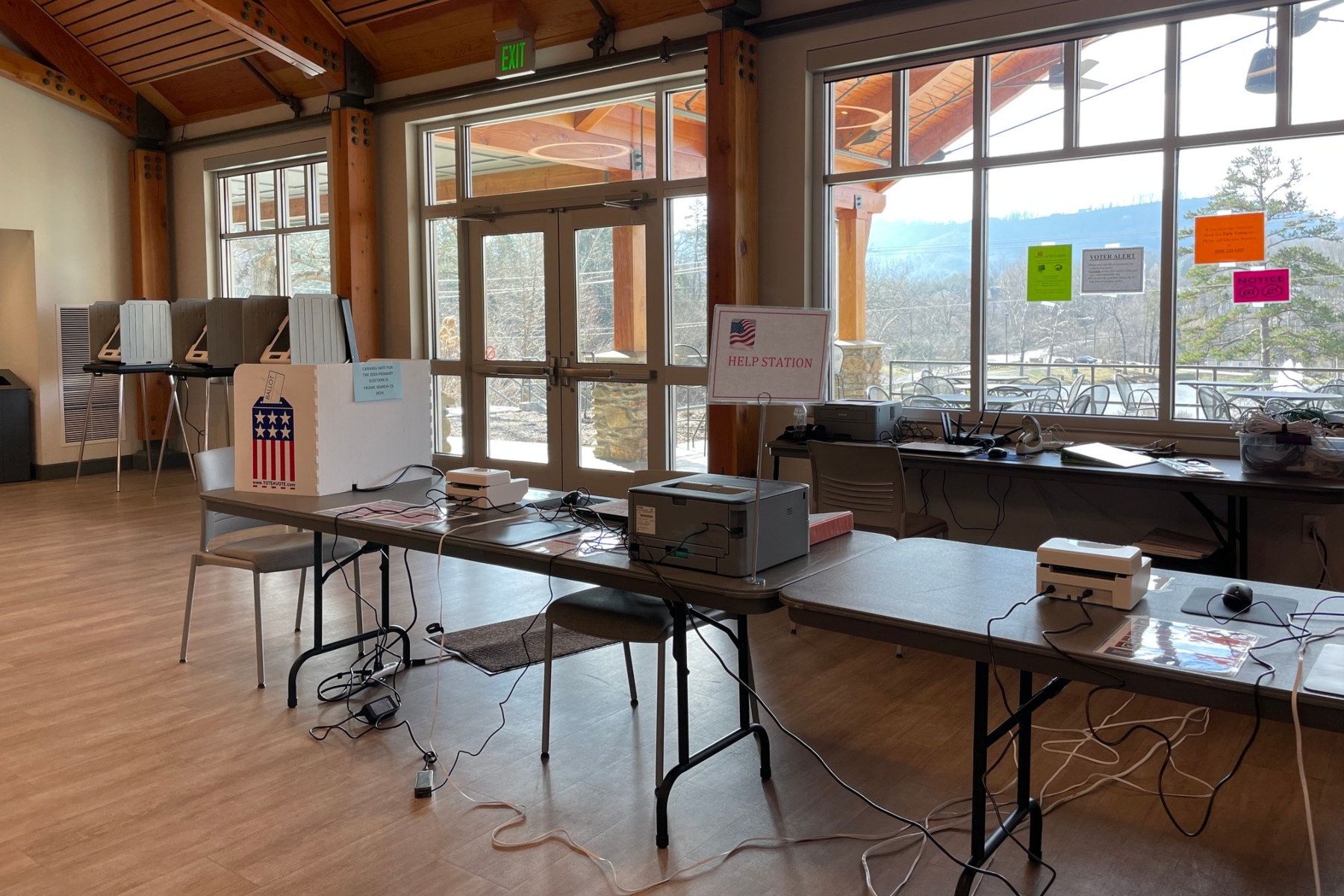 Election help station set up in a bright community room with voting equipment, a Help Station sign, and tangled cables on tables.