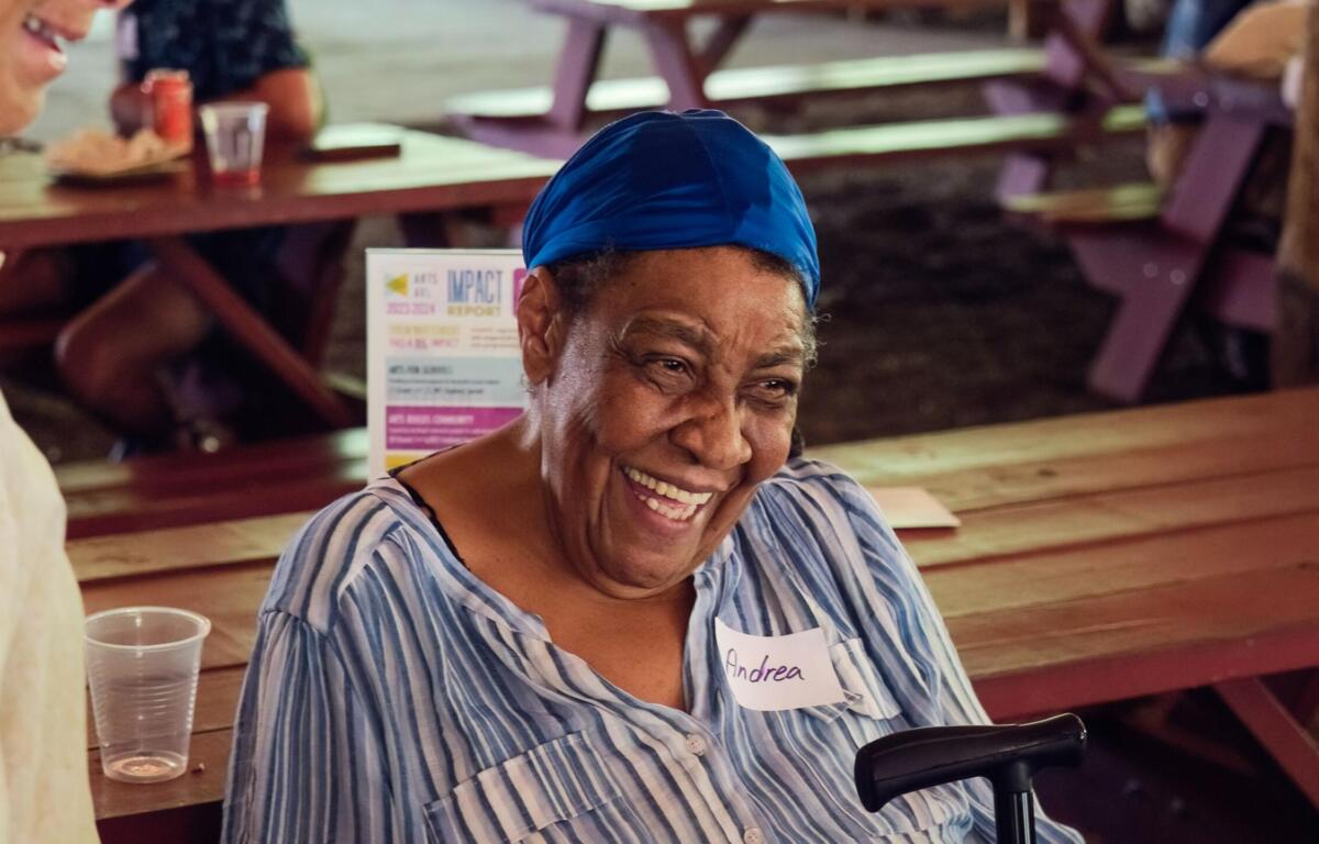 Smiling woman wearing a blue headscarf and striped shirt sits at a wooden picnic table; name tag reads Andrea.