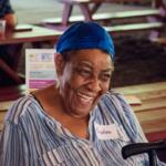 Smiling woman wearing a blue headscarf and striped shirt sits at a wooden picnic table; name tag reads Andrea.