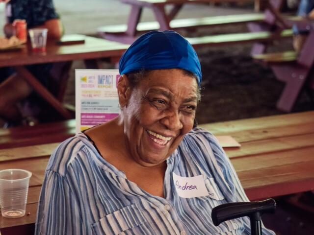 Smiling woman wearing a blue headscarf and striped shirt sits at a wooden picnic table; name tag reads Andrea.