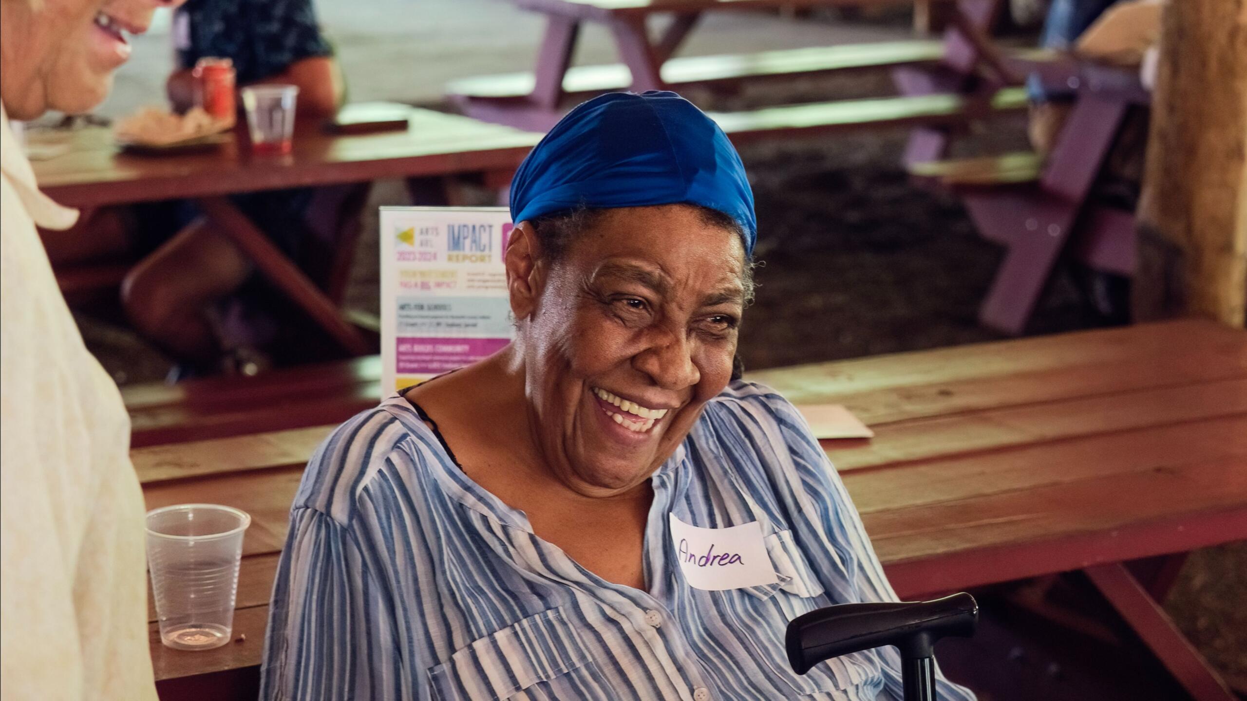 Smiling woman wearing a blue headscarf and striped shirt sits at a wooden picnic table; name tag reads Andrea.