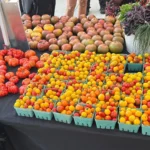 Assorted tomatoes in green trays and on the table at a market stall, with a white basket of herbs on the right and people in the background.