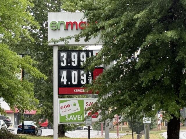Gas price sign partially obscured by trees, showing prices like 3.99 and 4.09 per litre.