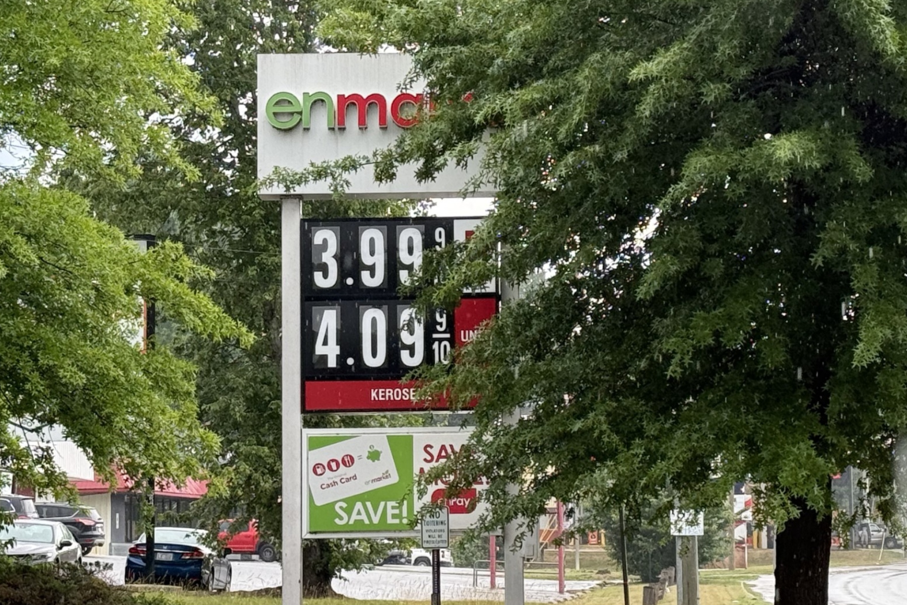 Gas price sign partially obscured by trees, showing prices like 3.99 and 4.09 per litre.