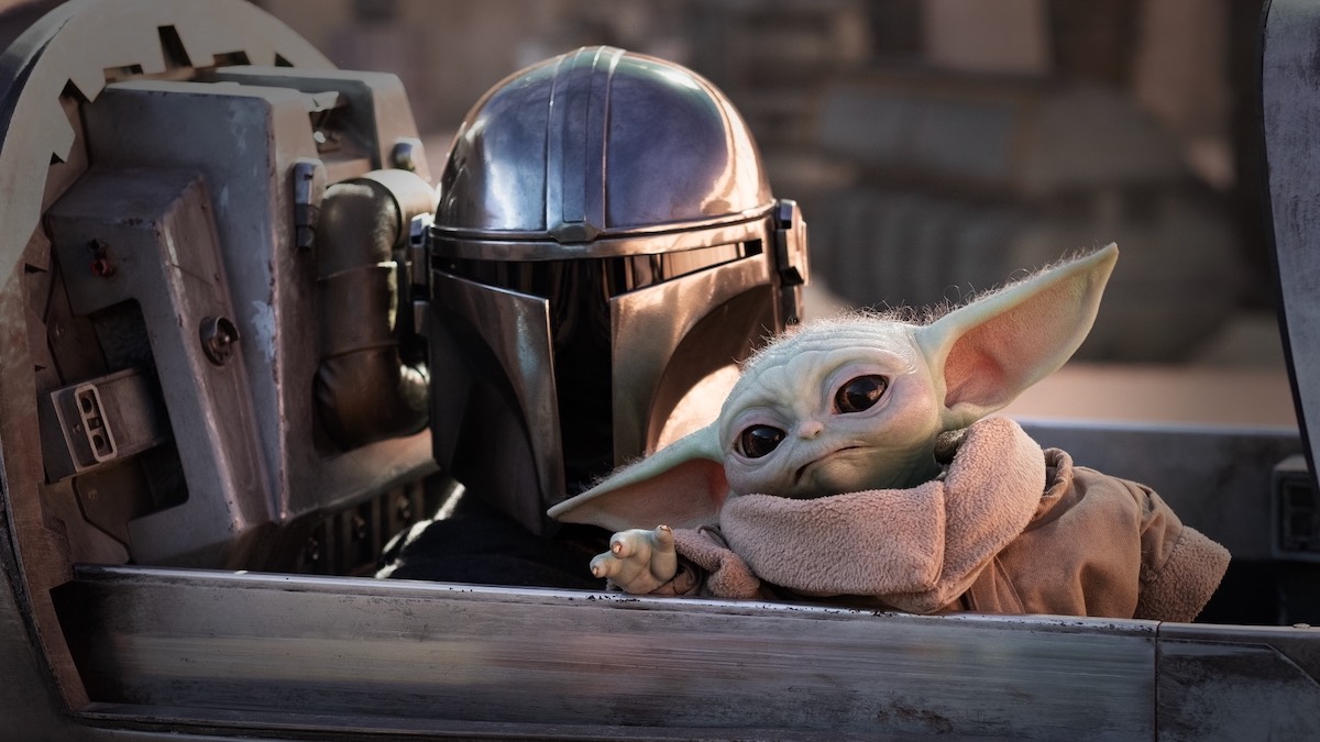 The Mandalorian sits beside Grogu in a sci‑fi vehicle, with Grogu leaning toward the camera edge.