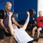 Man bench-presses heavy barbell with a spotter assisting in a gym with blue walls and wooden floor, while another man lifts dumbbells in the background.