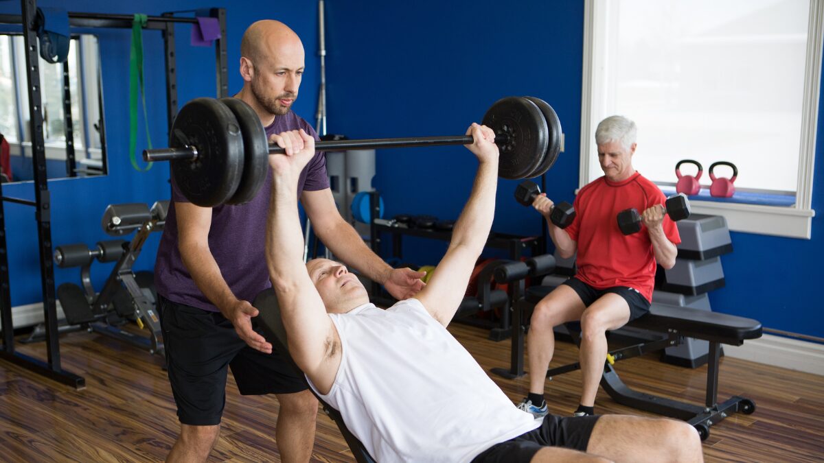Man bench-presses heavy barbell with a spotter assisting in a gym with blue walls and wooden floor, while another man lifts dumbbells in the background.