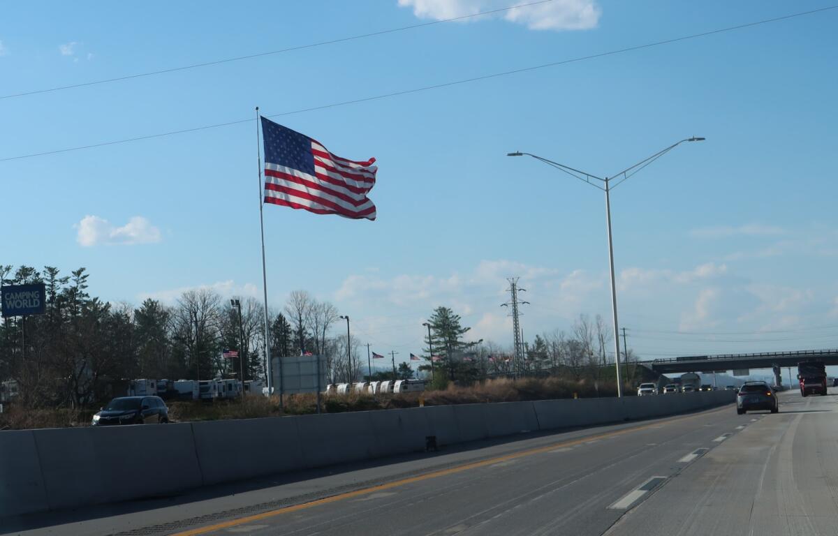 American flag waving on a tall flagpole along a highway with a clear blue sky and streetlights nearby.