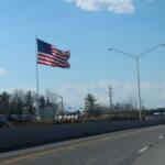American flag waving on a tall flagpole along a highway with a clear blue sky and streetlights nearby.