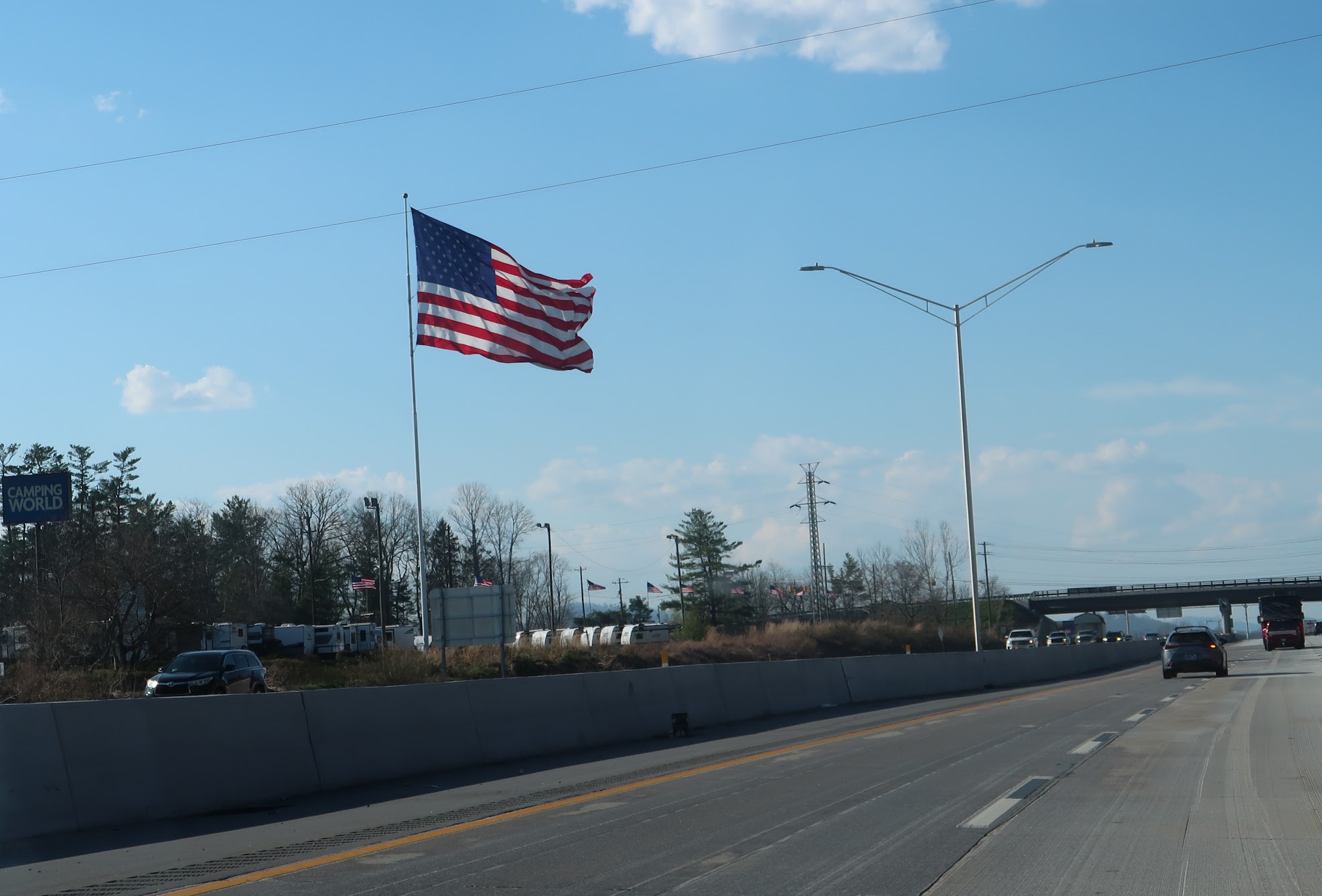 American flag waving on a tall flagpole along a highway with a clear blue sky and streetlights nearby.