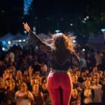 Performer seen from behind, raising arms on a night outdoor stage as a large cheering crowd watches.