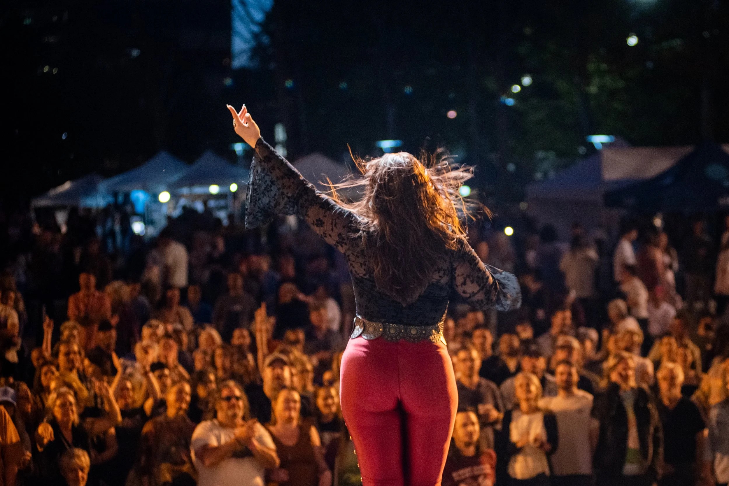 Performer seen from behind, raising arms on a night outdoor stage as a large cheering crowd watches.