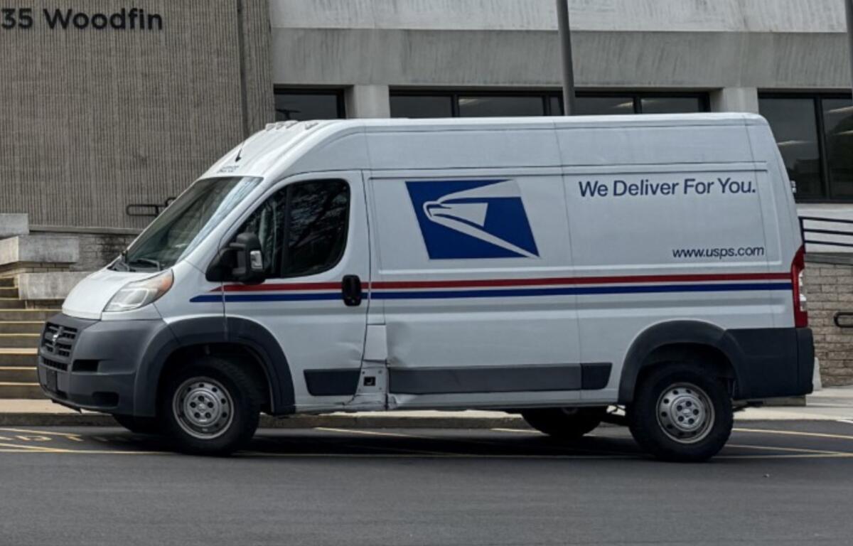 White USPS delivery van parked in front of a building, side text reads 'We Deliver For You' with USPS logo and website URL www.usps.com with red/blue stripes along the side.