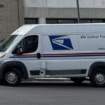 White USPS delivery van parked in front of a building, side text reads 'We Deliver For You' with USPS logo and website URL www.usps.com with red/blue stripes along the side.