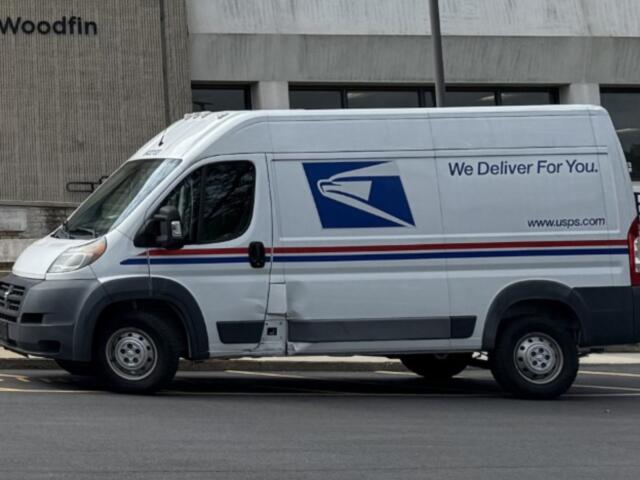 White USPS delivery van parked in front of a building, side text reads 'We Deliver For You' with USPS logo and website URL www.usps.com with red/blue stripes along the side.