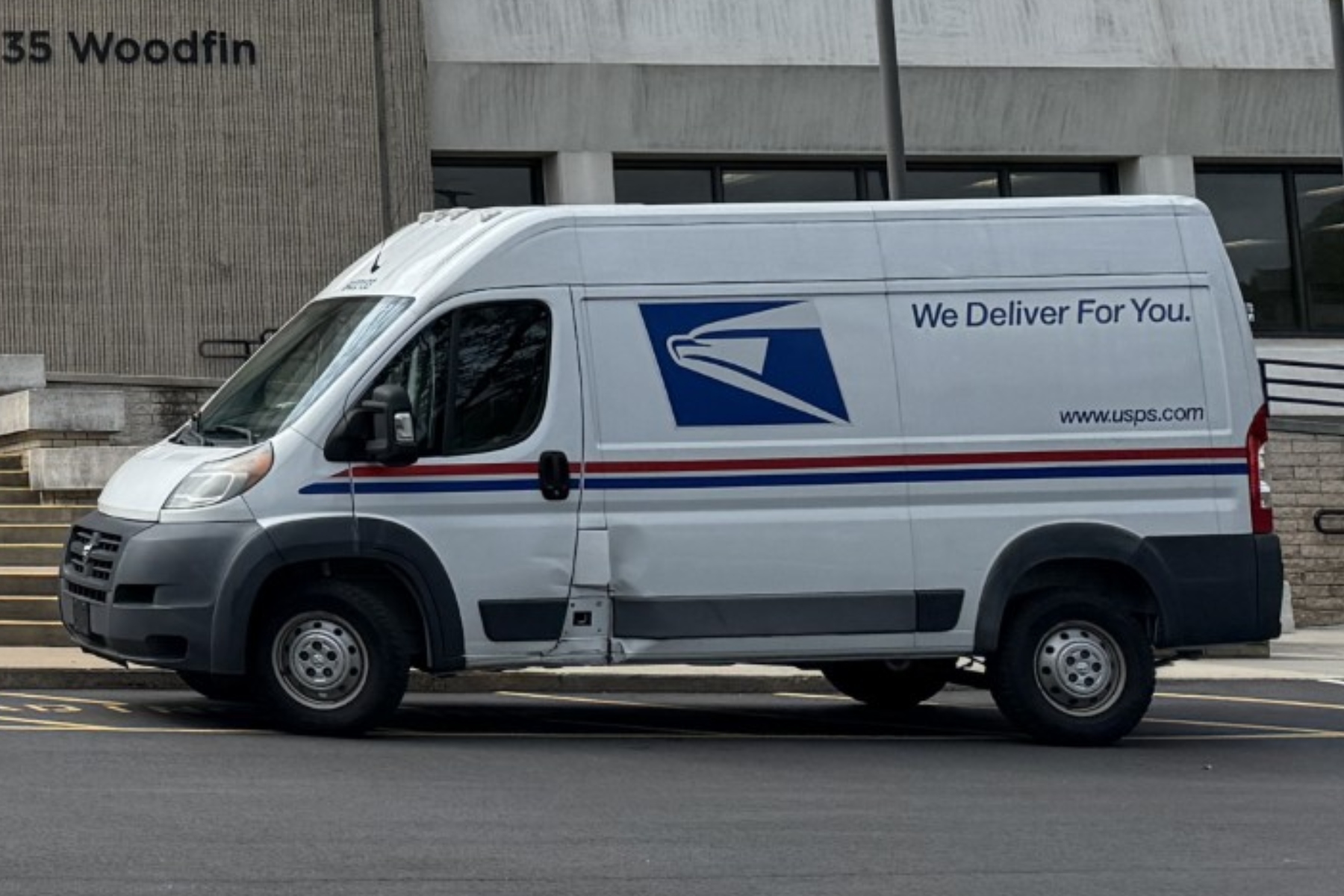 White USPS delivery van parked in front of a building, side text reads 'We Deliver For You' with USPS logo and website URL www.usps.com with red/blue stripes along the side.