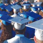 Back view of graduates in blue caps and gowns at a commencement ceremony, facing away from the camera.
