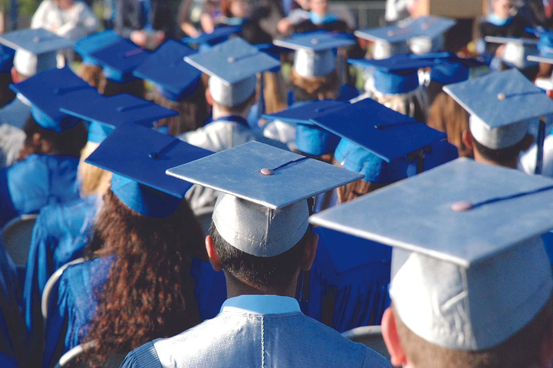 Back view of graduates in blue caps and gowns at a commencement ceremony, facing away from the camera.