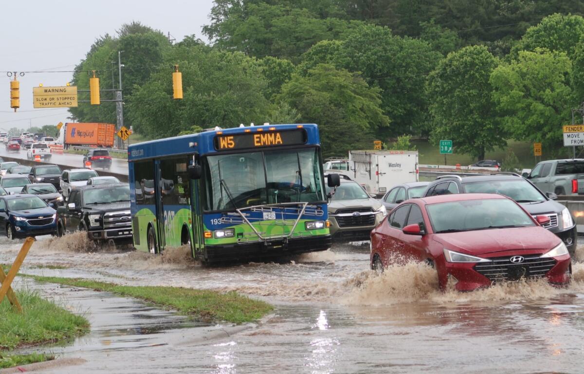 Blue city bus drives through a flooded street with cars splashing water around it, amid heavy traffic and green trees in background.