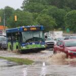 Blue city bus drives through a flooded street with cars splashing water around it, amid heavy traffic and green trees in background.