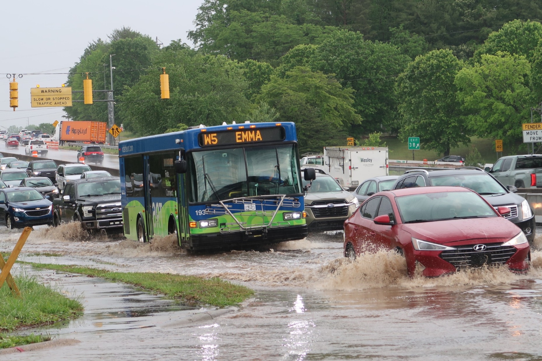 Blue city bus drives through a flooded street with cars splashing water around it, amid heavy traffic and green trees in background.