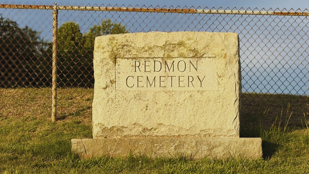 Stone cemetery marker engraved with REDMON CEMETERY, set on a raised base beside a chain-link fence and blue sky behind.