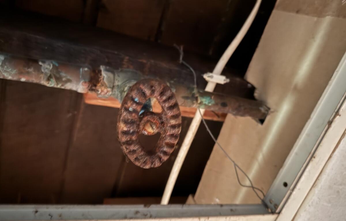 Rusty circular pulley hanging from a pipe in a dusty ceiling space.