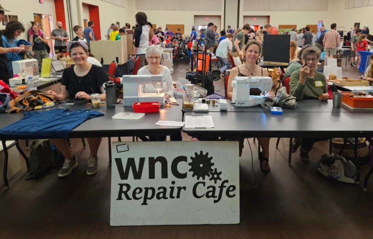 Group of people at a community repair cafe table with sewing machines, fabrics, and tools; the banner reads "WNC Repair Cafe" in the foreground.