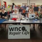 Group of people at a community repair cafe table with sewing machines, fabrics, and tools; the banner reads "WNC Repair Cafe" in the foreground.