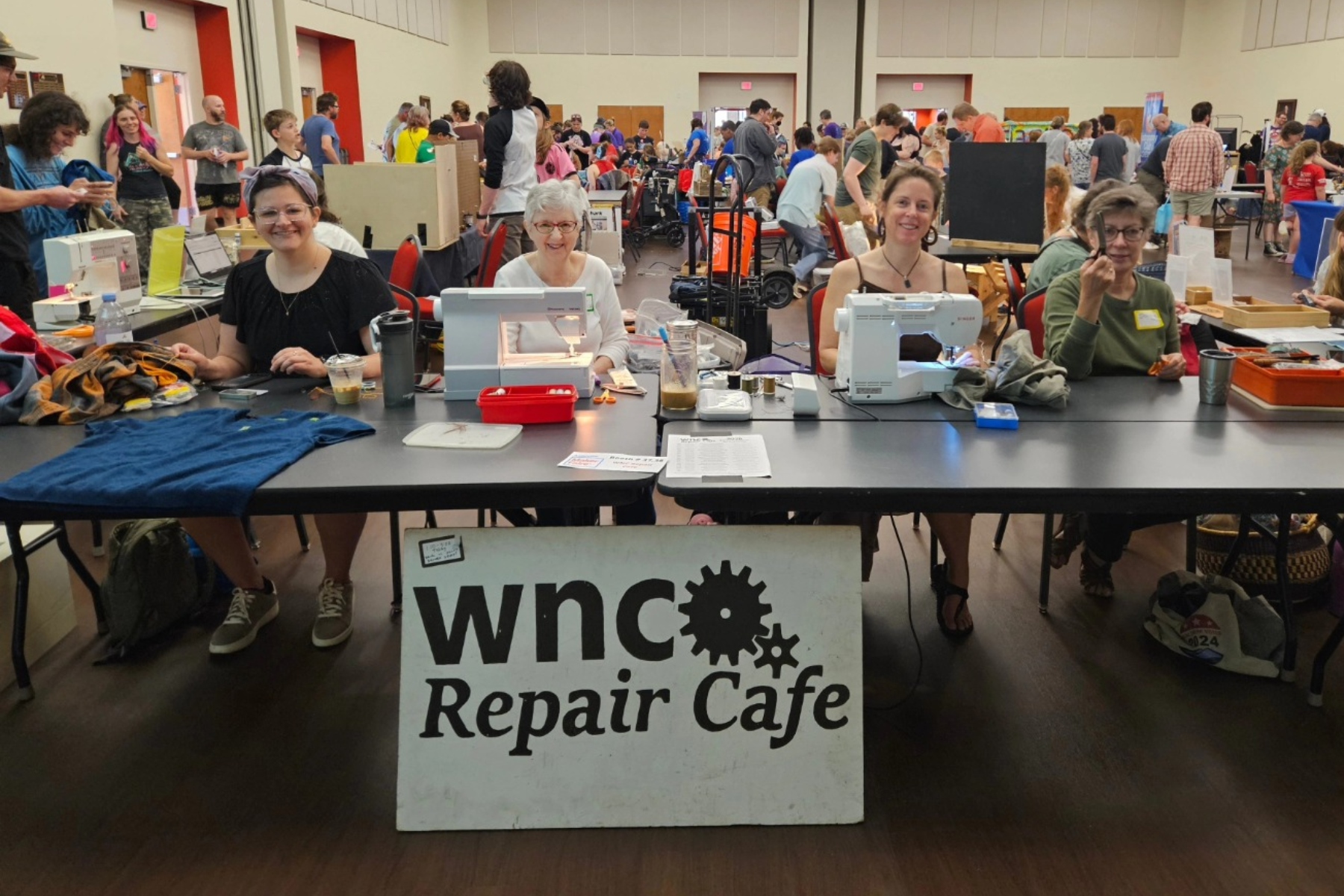 Group of people at a community repair cafe table with sewing machines, fabrics, and tools; the banner reads "WNC Repair Cafe" in the foreground.