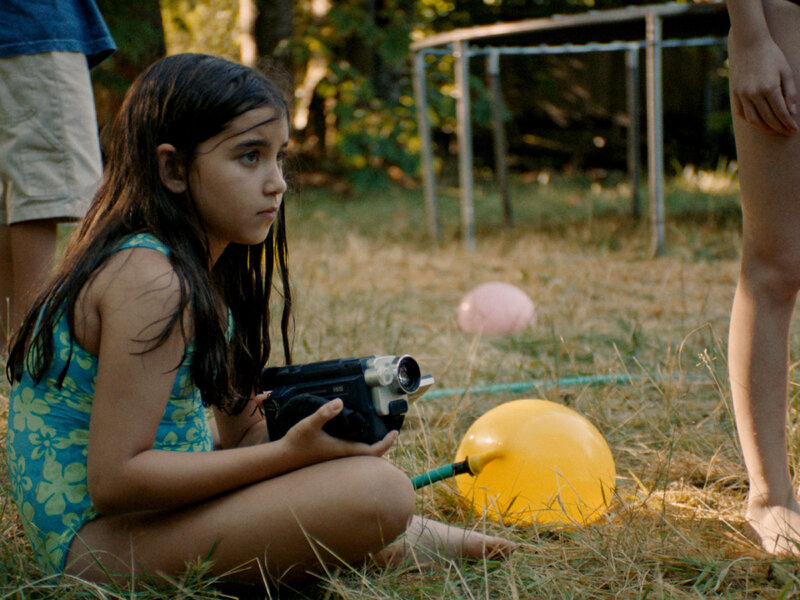 Young girl sitting on grass outdoors, holding a video camera and watching something off to the side.