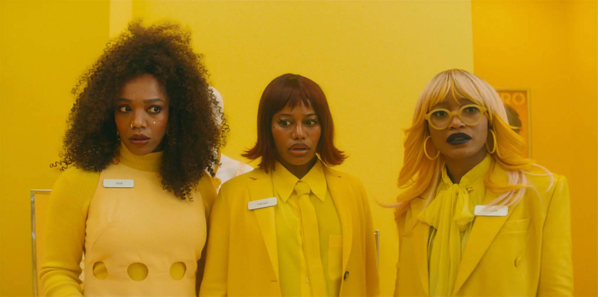 Three women in yellow outfits stand together and look off-camera in a bright yellow setting.