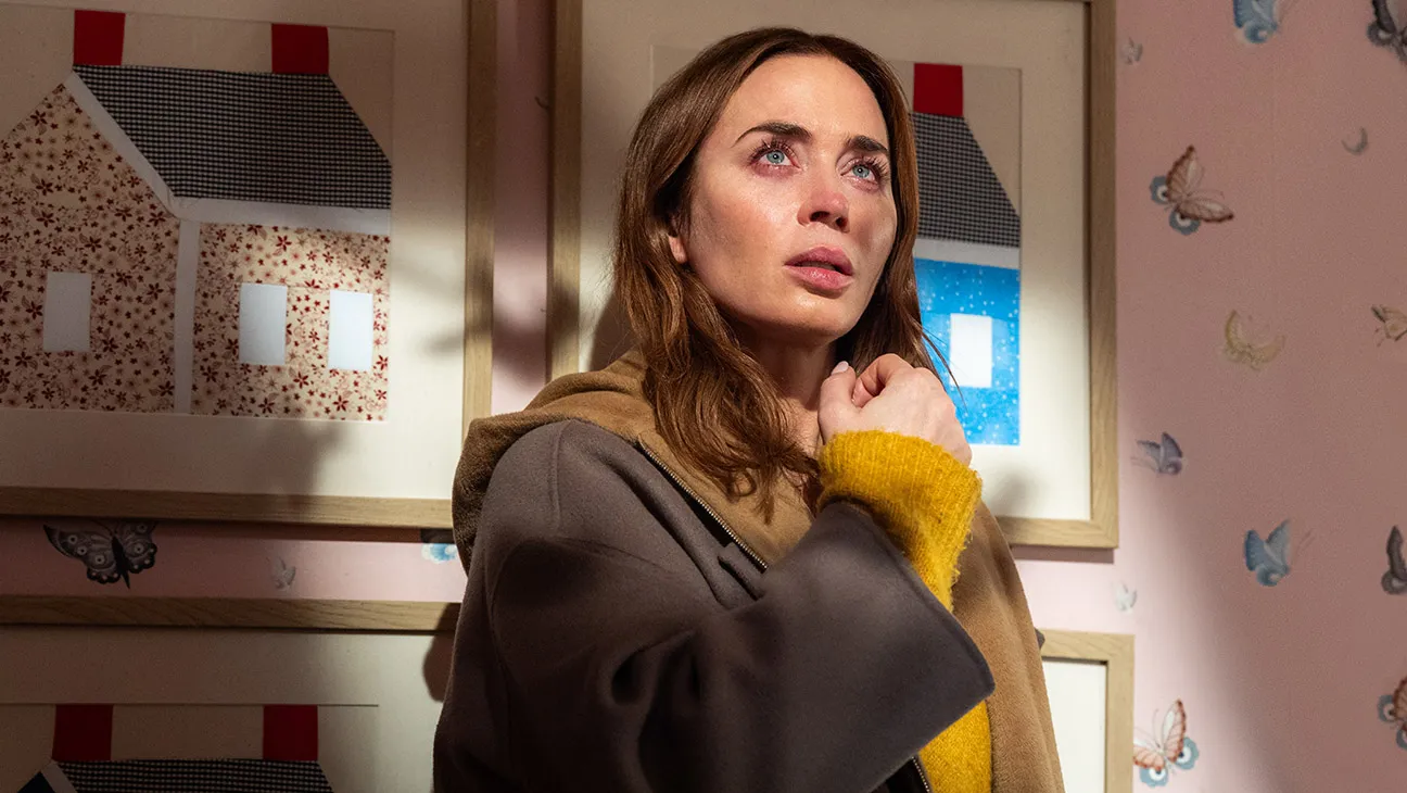 Woman with brown hair in a tan coat and yellow sweater, standing in an art gallery and looking up with a concerned expression.
