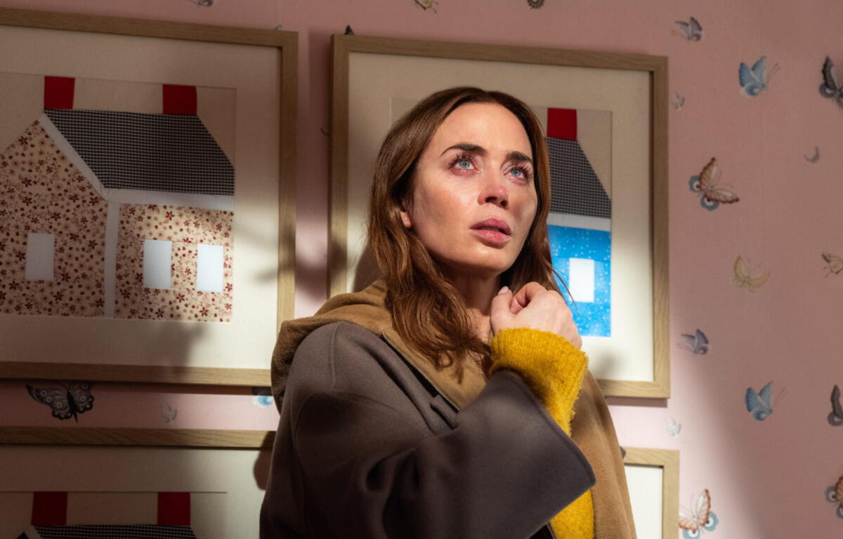 Woman with brown hair in a tan coat and mustard-yellow sweater looks upward, standing in a pink room with framed artwork behind her.