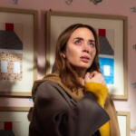 Woman with brown hair in a tan coat and mustard-yellow sweater looks upward, standing in a pink room with framed artwork behind her.