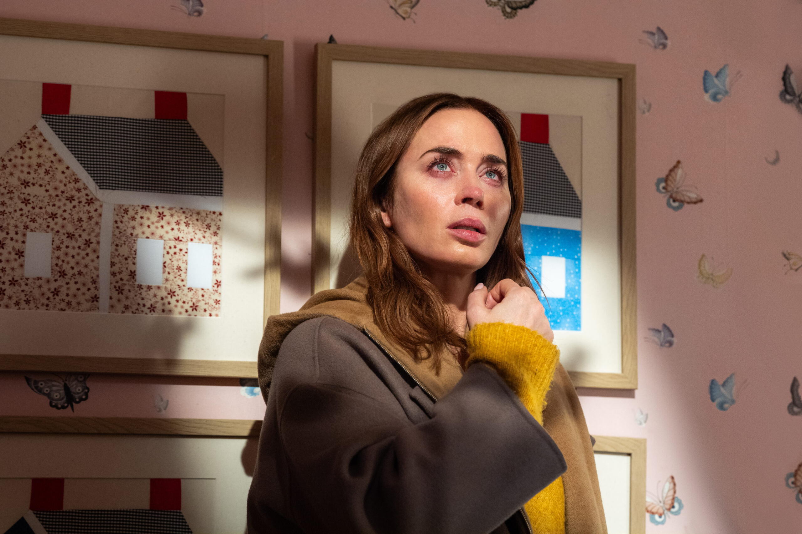 Woman with brown hair in a tan coat and mustard-yellow sweater looks upward, standing in a pink room with framed artwork behind her.