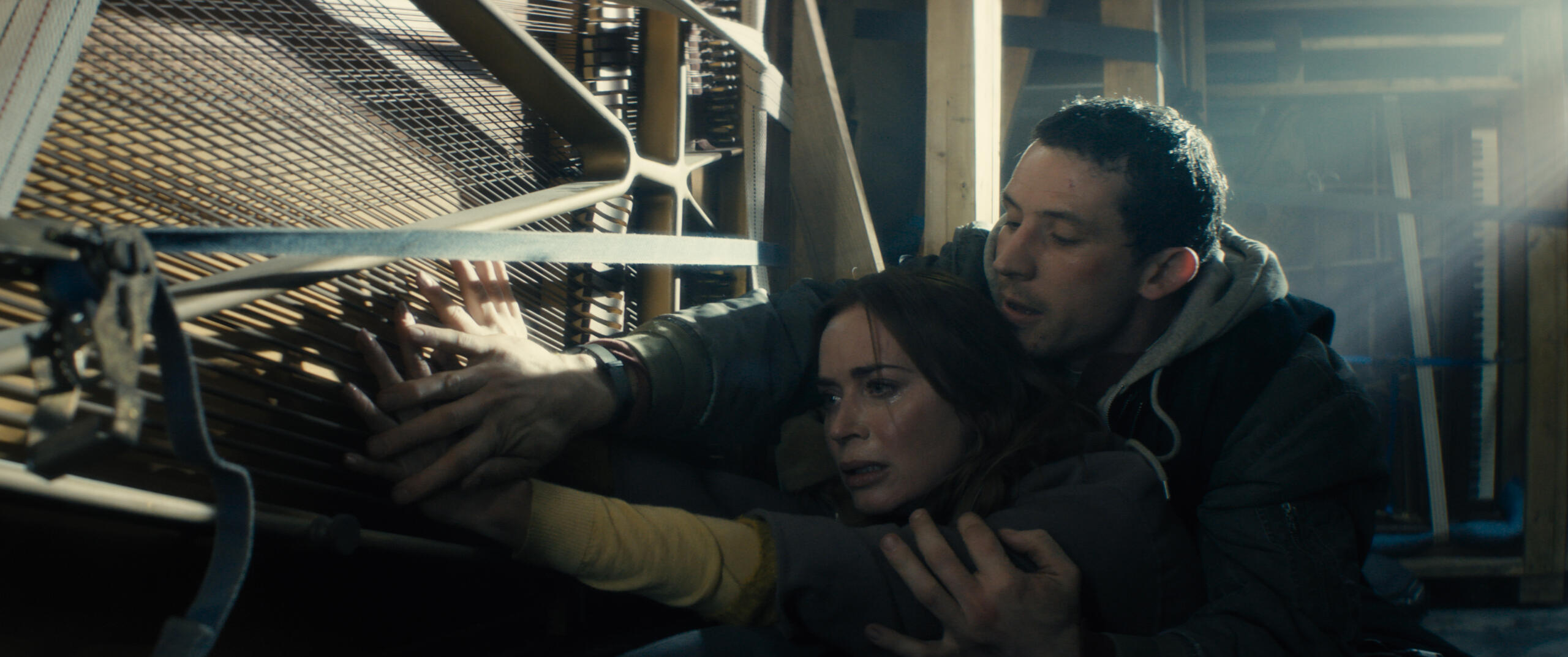 Woman crying as a man shields her, both clinging to a large piano in a storage-like space