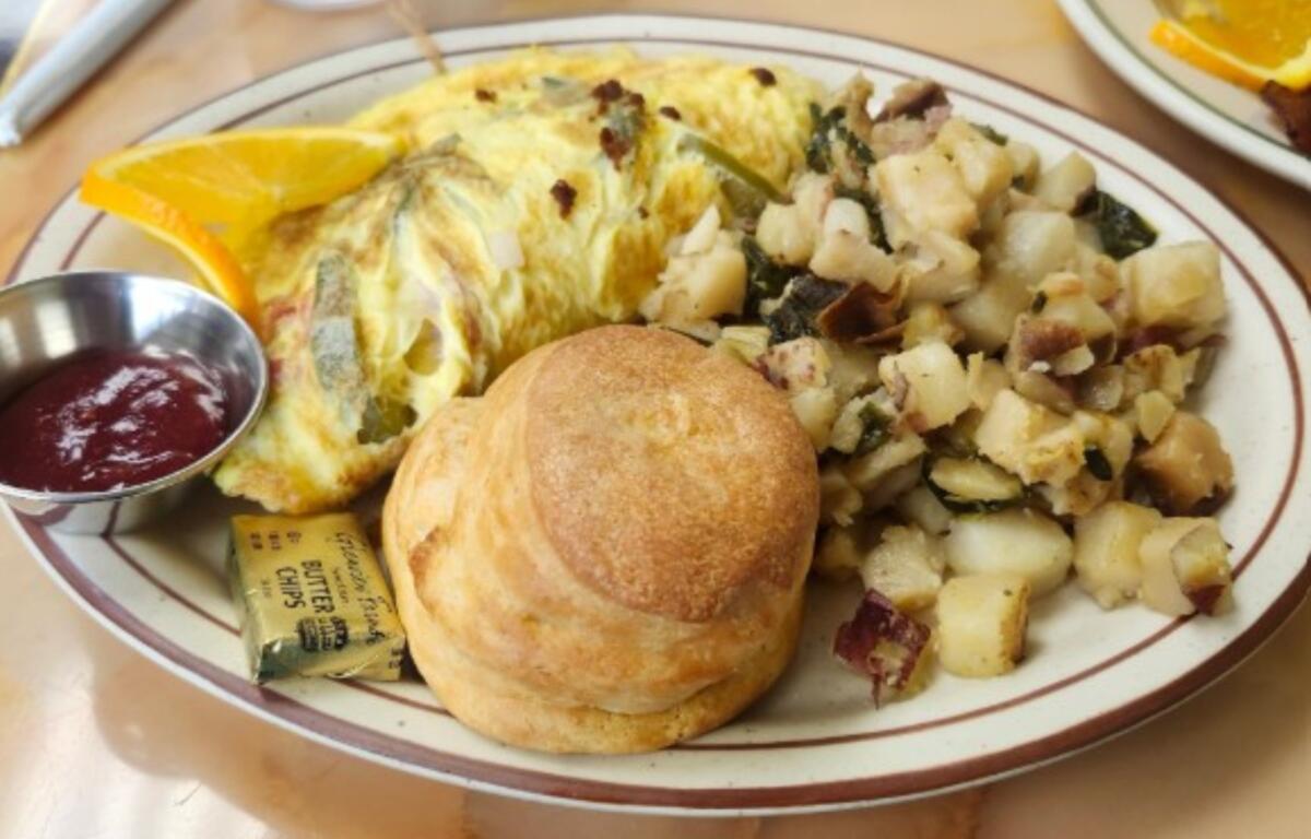 Breakfast plate with scrambled omelette, hash browns, a biscuit, orange wedge, and a cup of jam on the side.