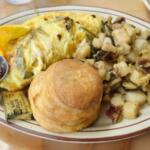Breakfast plate with scrambled omelette, hash browns, a biscuit, orange wedge, and a cup of jam on the side.