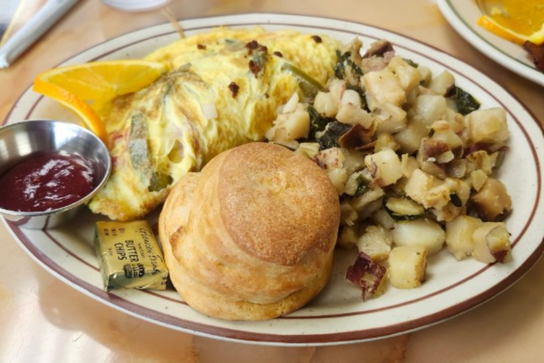 Breakfast plate with scrambled omelette, hash browns, a biscuit, orange wedge, and a cup of jam on the side.