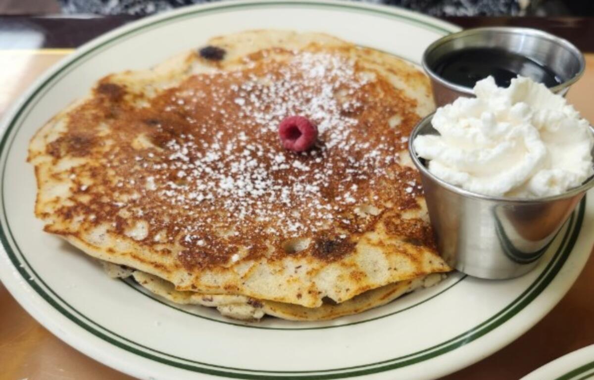 Abeja's House Cafe's gluten-free blueberry pancakes, dusted with powdered sugar, topped with whipped cream, and paired with maple syrup and a side of bacon.
